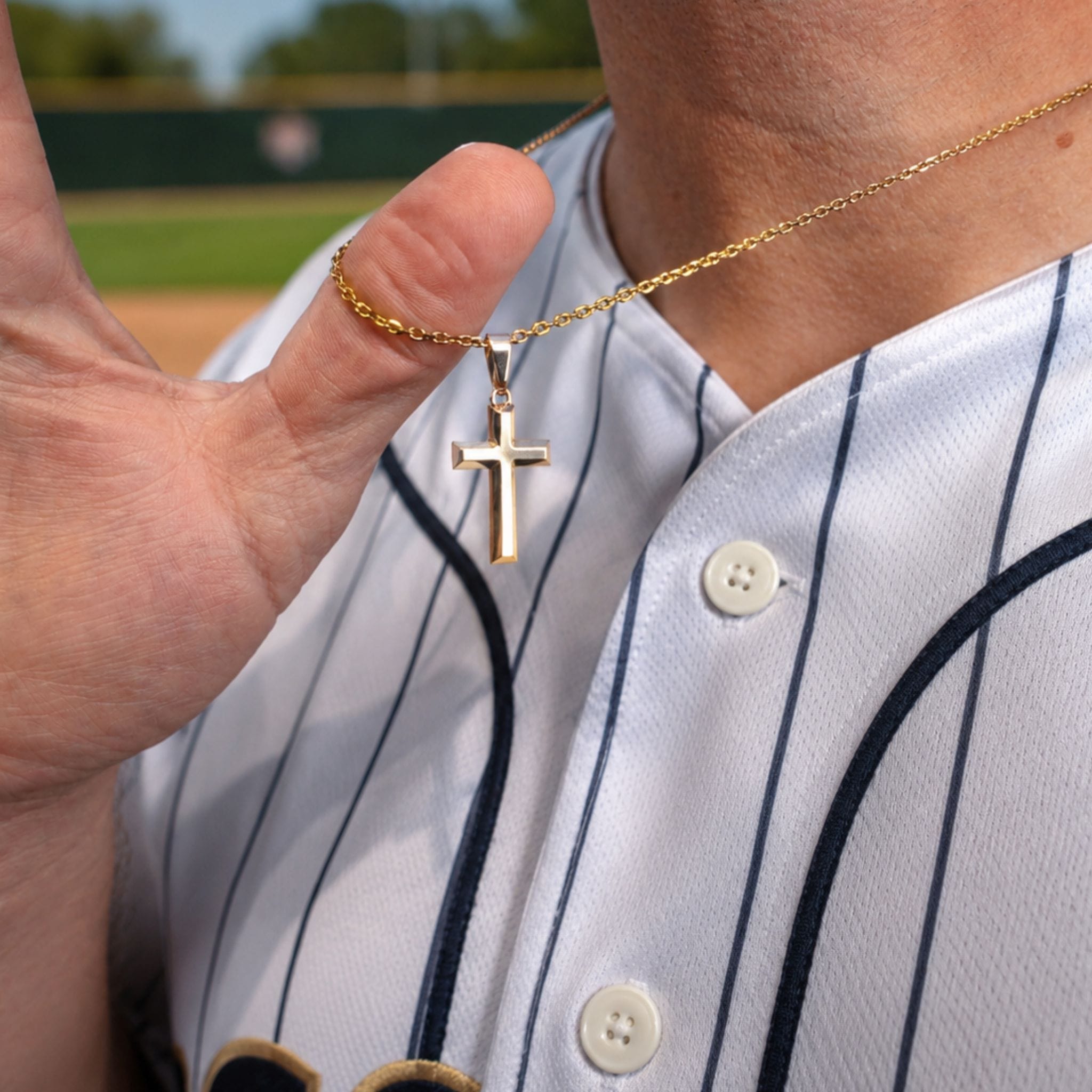 A gold cross pendant necklace worn by a baseball player in a white pinstripe jersey, with the player's hand holding the chain to show the scale of the pendant.