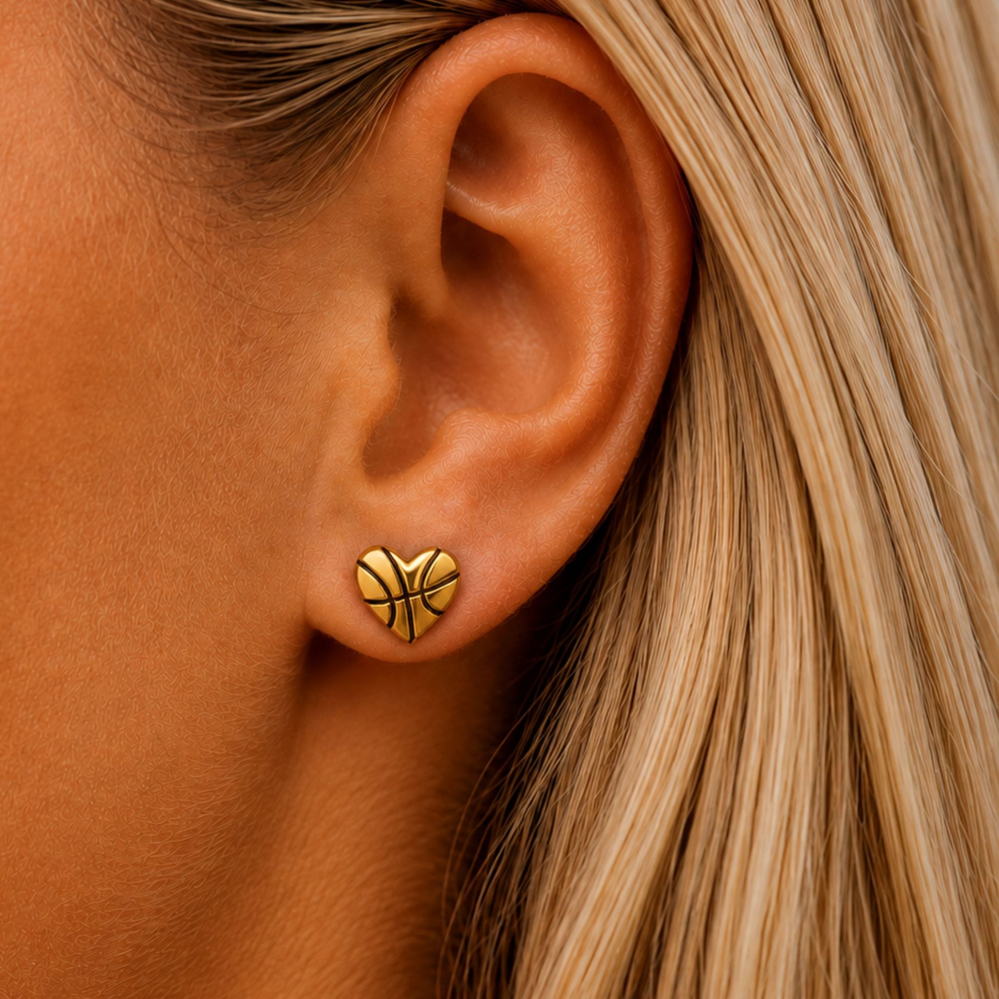 A close-up of a woman's ear wearing a small gold heart-shaped stud earring with a basketball design, next to her blonde hair.