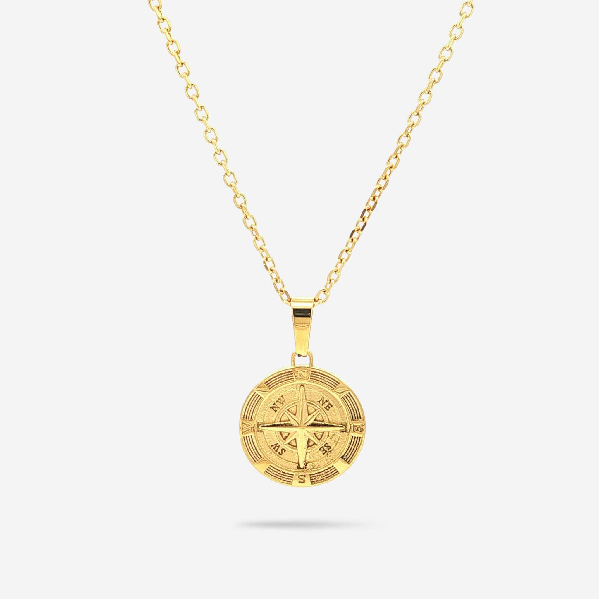 A gold pendant necklace featuring a detailed compass charm, suspended from a gold cable chain against a light gray background.