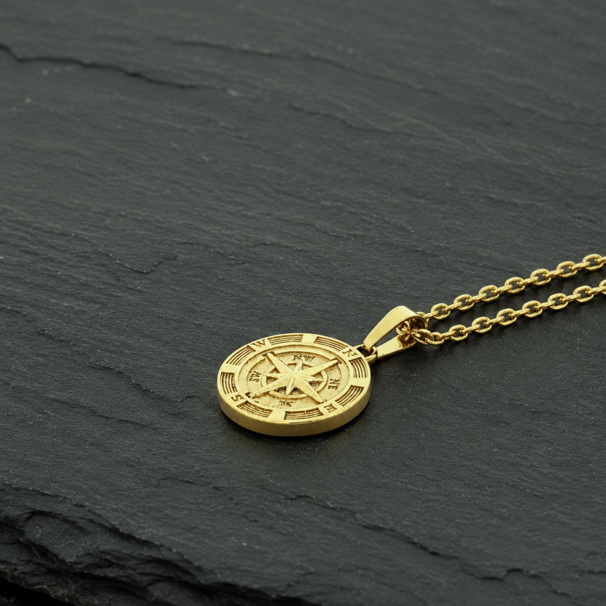 A gold-toned compass pendant necklace resting on a textured dark gray slate surface.