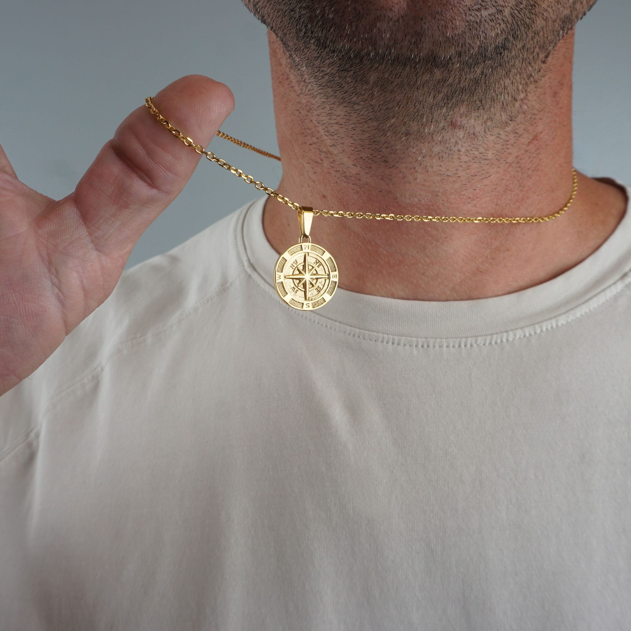 A close-up of a man's neck and chest wearing a gold chain necklace with a round compass pendant, while his hand holds a section of the chain against a plain gray background.