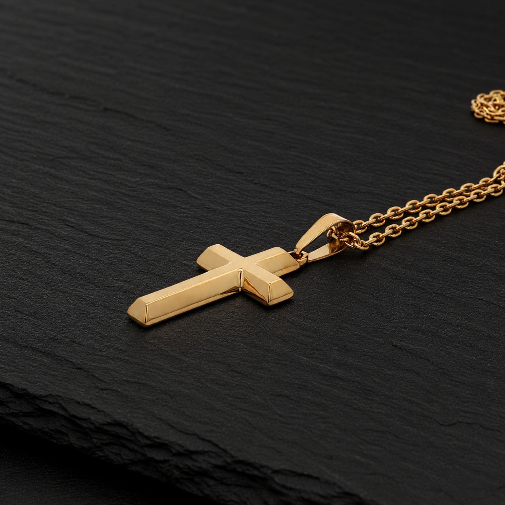 A gold cross pendant necklace with a matching chain, displayed on a textured black slate surface.