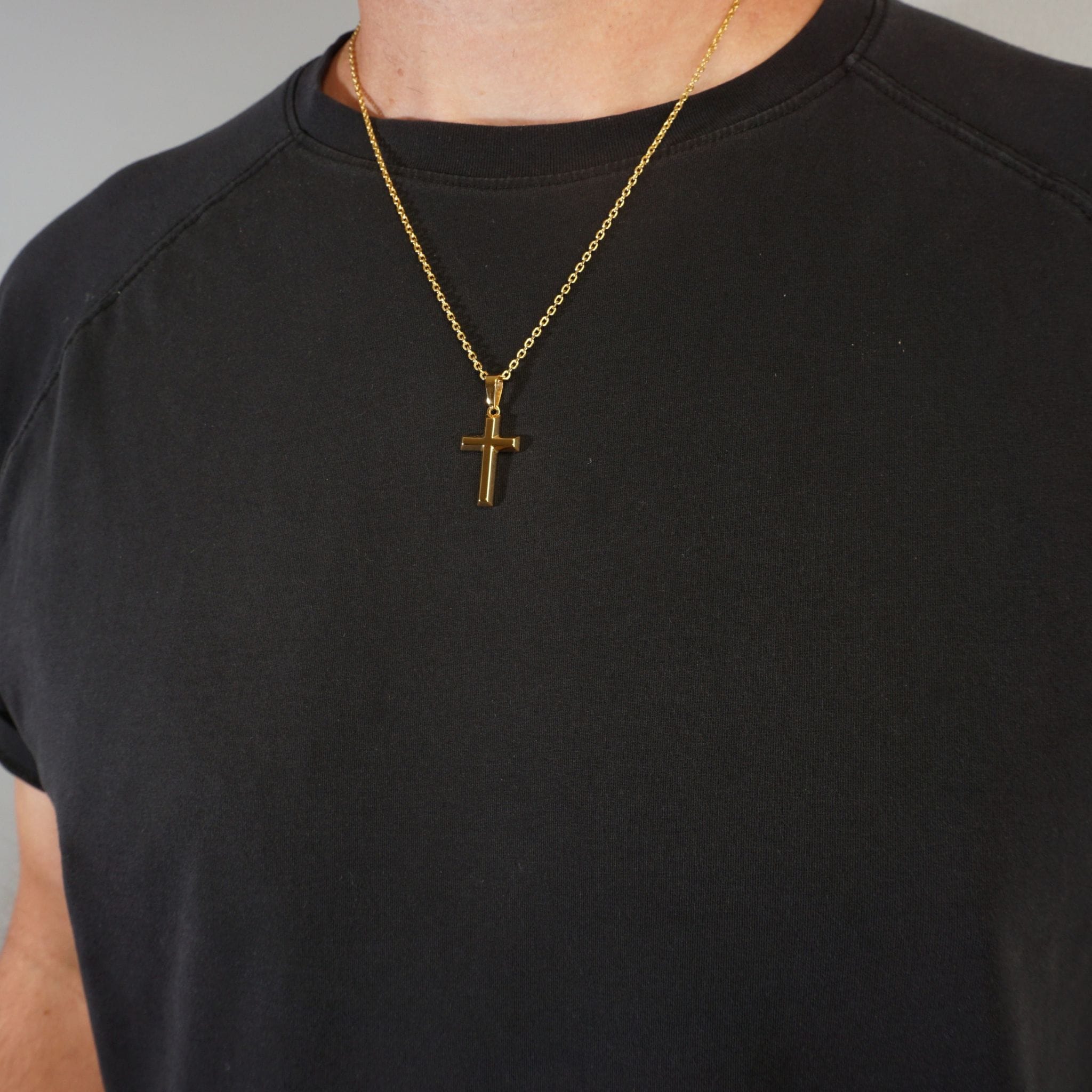 A close-up of a person wearing a gold cross pendant necklace on a black crew-neck t-shirt against a neutral gray background.
