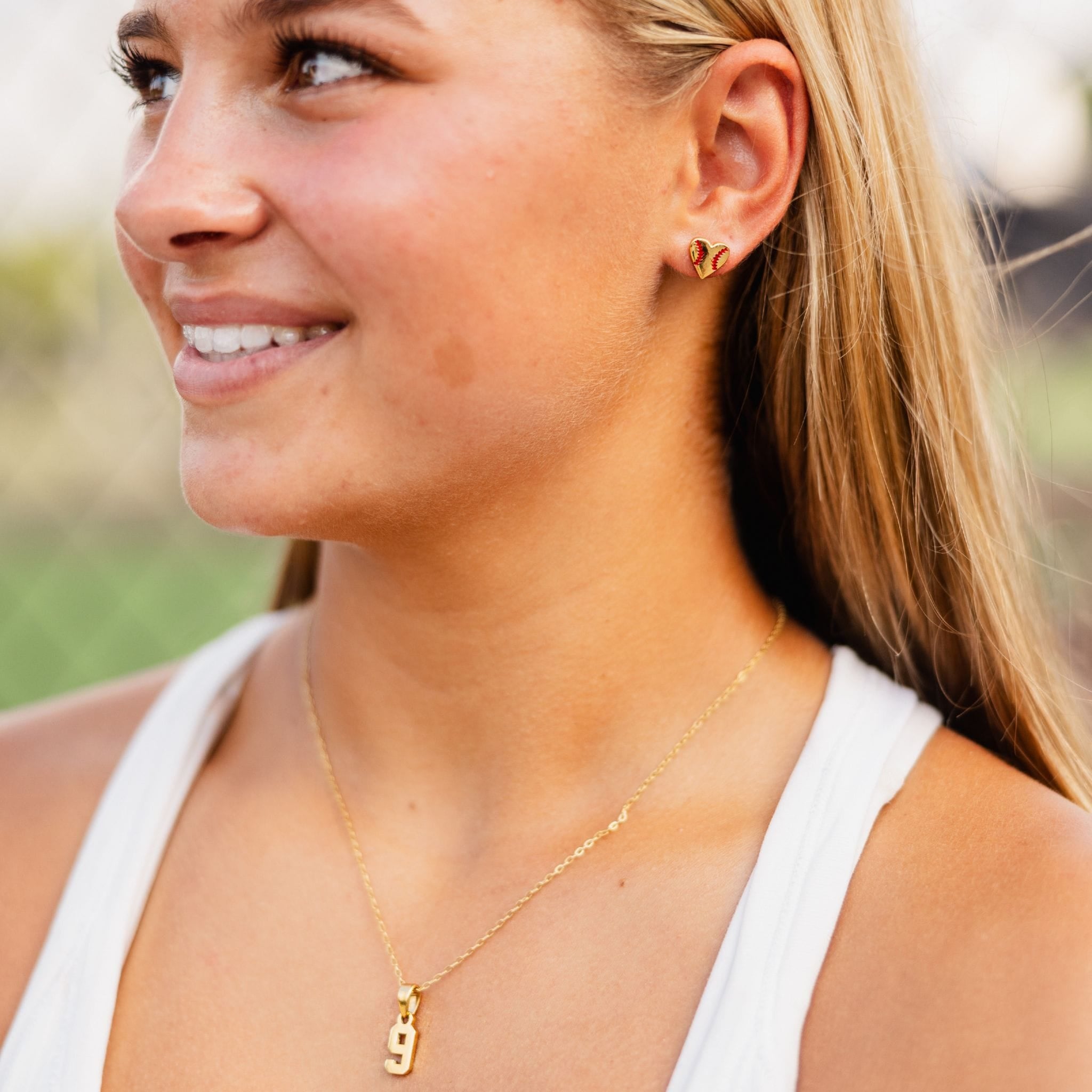 A lifestyle shot of a smiling woman wearing a number necklace with a number 9 pendant and a matching gold heart shaped baseball earrings