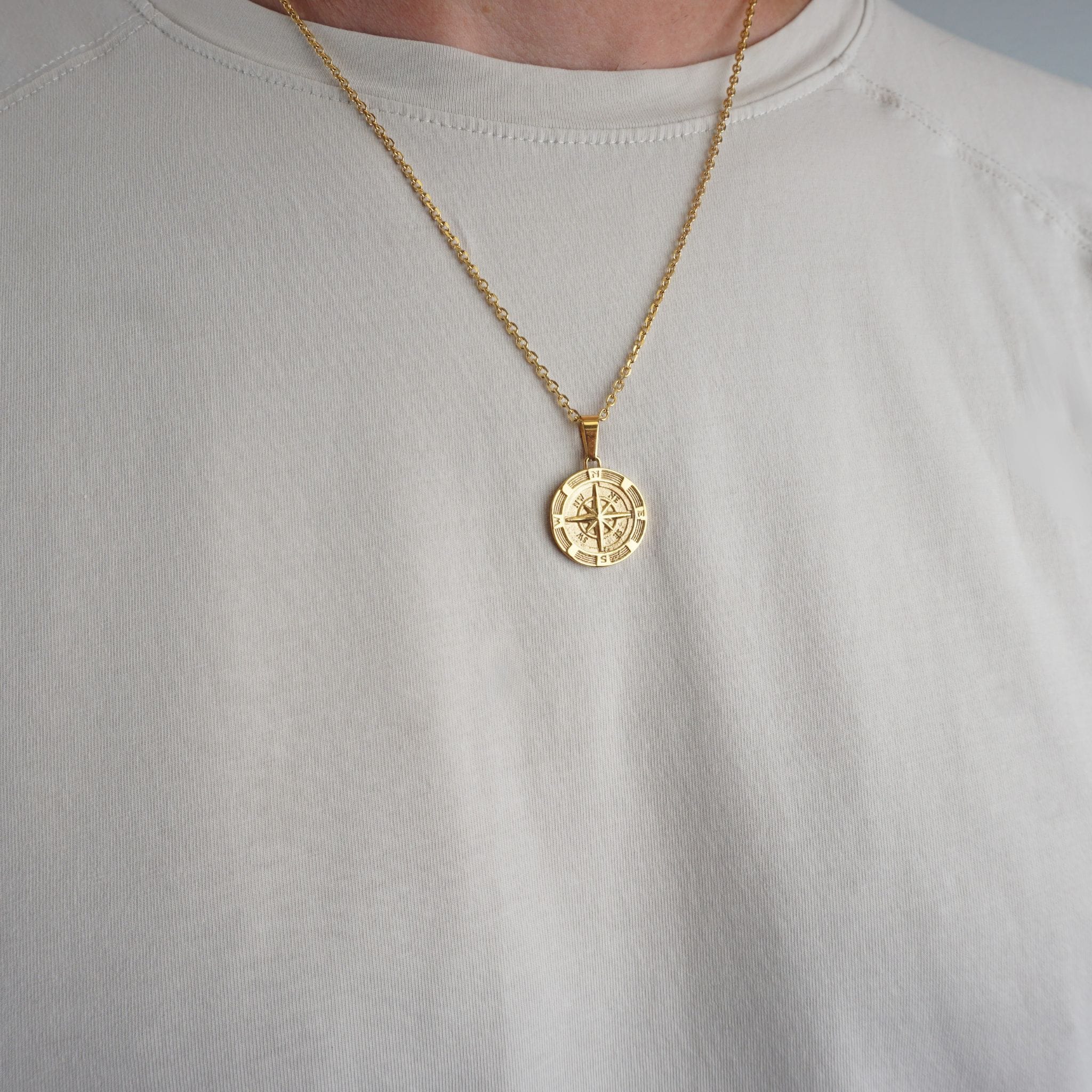 A gold necklace with a circular compass pendant is worn by a person wearing a light beige crewneck t-shirt.