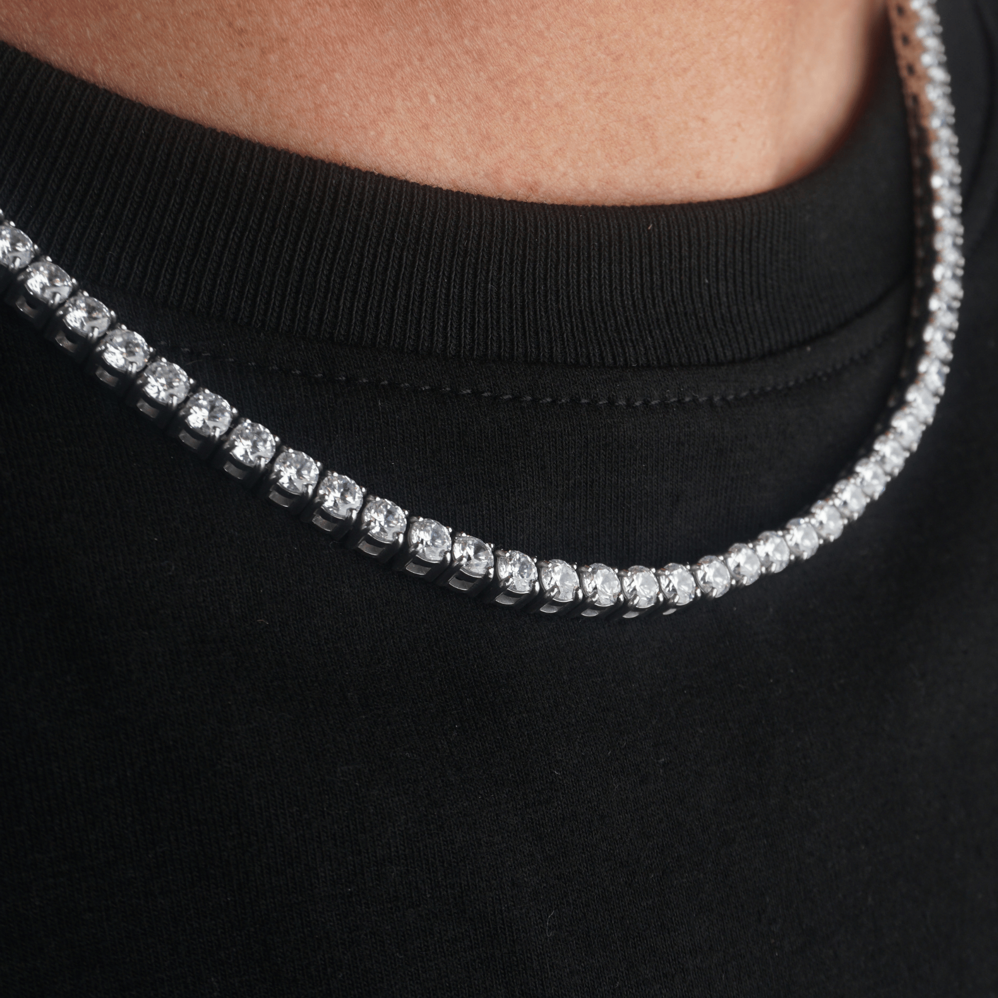 A close-up of a silver-toned tennis chain necklace with clear cubic zirconia stones worn around a person's neck over a black crewneck shirt.