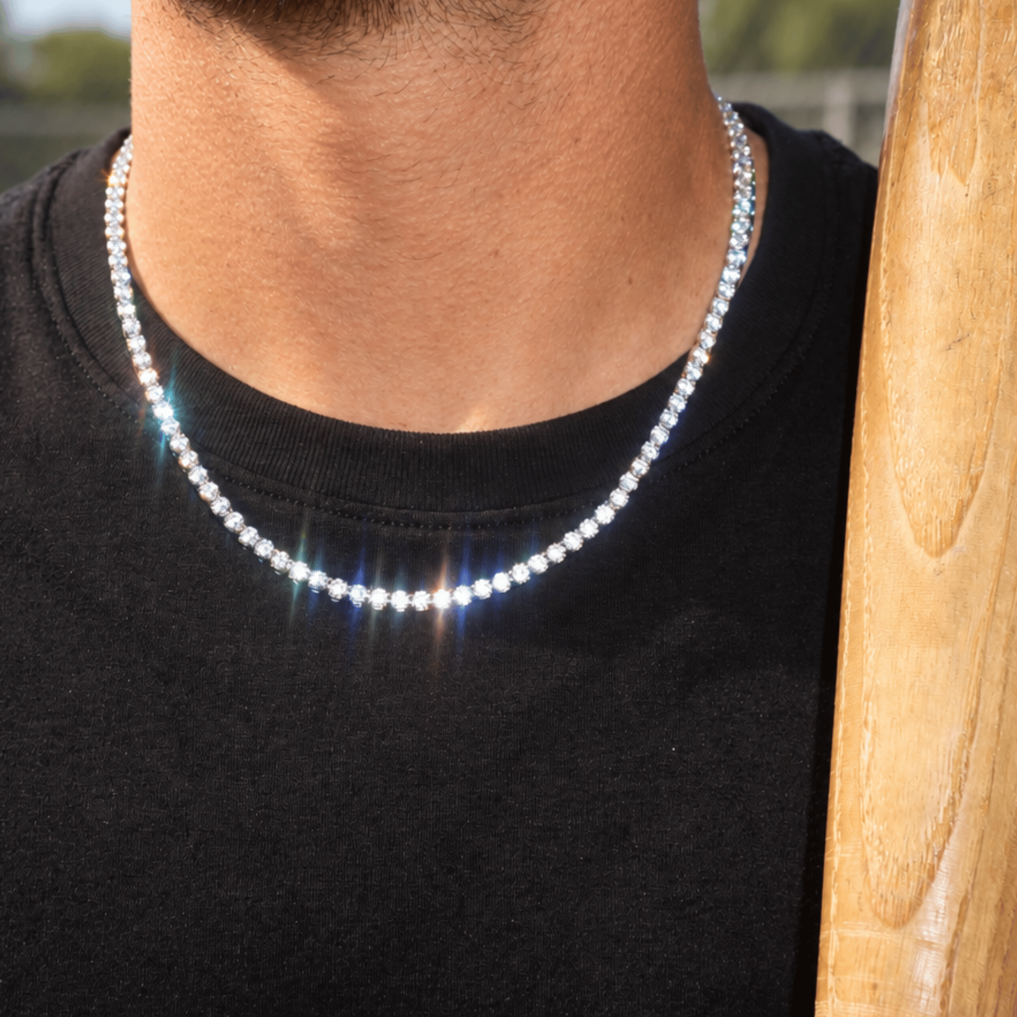 A close-up of a person wearing a sparkling silver tennis chain necklace over a black t-shirt, with a wooden bat visible on the right side.